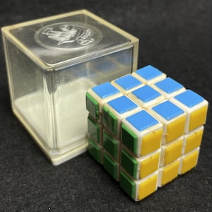 Op de afbeelding: Een klassieke Rubik's Cube met blauwe, gele, groene en witte vierkanten. De puzzel staat naast een doorzichtige plastic doos met een zilveren embleem op het deksel. Een populair hersenkraker.