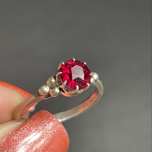 May include: A silver ring featuring a large, faceted, round, red gemstone. The ring has a delicate band and small decorative beads on either side of the gemstone. The ring is held against a gray background.
