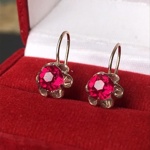 May include: A pair of gold-coloured earrings with a flower design. Each earring has a vibrant, faceted, red gemstone at the centre, surrounded by delicate petals. The earrings are displayed in a red jewellery box.