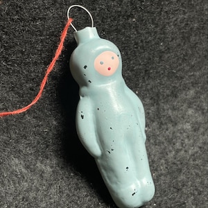 以下が含まれることがあります： 水色の、ヴィンテージスタイルの人物型クリスマスオーナメント。オーナメントは、目に小さな黒い点、口に赤い点があるピンク色の顔をしています。吊り下げ用の銀色のワイヤーと赤い紐が付いています。