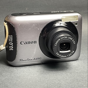 May include: Silver Canon PowerShot A490 digital camera with a black lens and flash. The camera has a 10.0 megapixel resolution and a 3.3x zoom lens. The camera is compact and has a wrist strap.