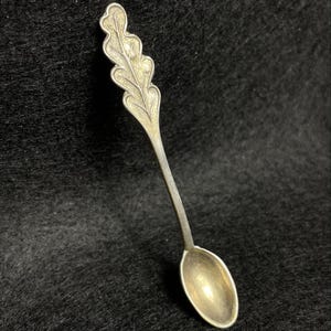 Può includere: Un cucchiaio d'argento con un design a foglia di quercia sul manico. Il cucchiaio ha una piccola coppa ovale e un manico lungo e sottile. Il design della foglia di quercia è dettagliato e aggiunge un elemento decorativo.