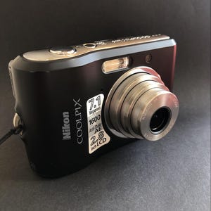 May include: Black Nikon Coolpix digital camera with a silver lens and flash. The camera has a 2.8-inch LCD screen and features 7.1 megapixels and 1600 ISO. The camera has a black strap attached to the side.