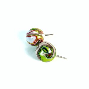 Pode incluir: Dois brincos redondos com um padrão em espiral em branco, verde, vermelho e castanho. Os brincos têm um pino prateado. Os brincos estão sobre um fundo branco.