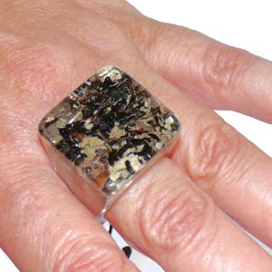 May include: A square-shaped ring featuring a clear, resin-like top surface that encases a textured, multi-colored inlay of black, tan, and metallic gold fragments. The band of the ring is smooth and silver-toned, providing a clean, reflective finish.