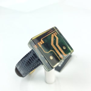 May include: A black leather bracelet with a clear resin square charm featuring a green circuit board with copper traces.
