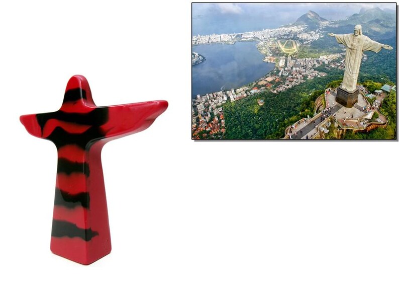 May include: A red and black abstract sculpture of a figure with outstretched arms, resembling the Christ the Redeemer statue. The background shows an aerial view of Rio de Janeiro, Brazil, with the Christ the Redeemer statue on a hilltop.