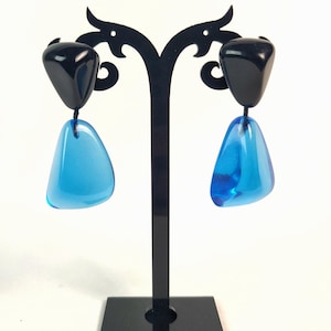 Op de afbeelding: Een paar geometrische oorbellen op een zwarte displaystandaard. Elke oorbel heeft een zwarte, driehoekige bovenkant en een doorschijnende, blauwe, druppelvormige onderkant. De oorbellen worden gepresenteerd tegen een witte achtergrond.