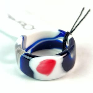 May include: A smooth, circular ring crafted from resin or glass, featuring a marbled pattern of deep blue, opaque white, and a single prominent red spot. A thin black string is looped through the center of the band, which is set against a white background.