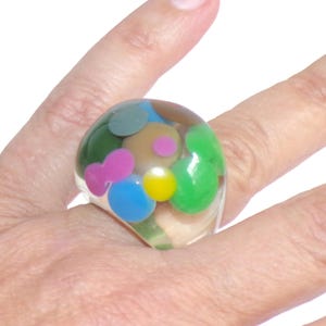 May include: A clear resin ring with colorful, rounded shapes in green, blue, pink, yellow, and brown. The ring is a chunky, statement piece with a playful, artistic design. The ring is worn on a finger.