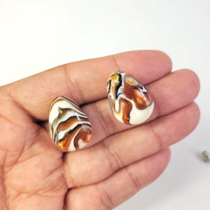 May include: Two teardrop-shaped beads with a marbled design. The beads feature a mix of white, brown, and black swirls. The beads are smooth and glossy, with a polished finish. The beads are held in a hand.