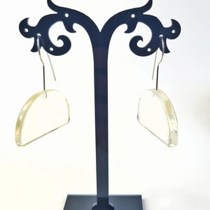 May include: A pair of clear, semi-circular earrings with silver-colored hooks. The earrings are displayed on a black stand with a decorative top, against a white background. The earrings have a slight yellow tint.