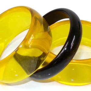 May include: Three translucent bangles. Two are a bright yellow, and one is black. The yellow bangles have a slightly rounded shape, while the black bangle is smooth and glossy. The bangles are stacked together.
