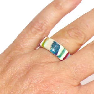 Może przedstawiać: Kolorowy pierścionek o prostokątnym kształcie. Pierścionek zawiera mieszankę kolorów, w tym biały, niebieski, zielony, fioletowy i czerwony. Pierścionek jest noszony na palcu, prezentując swój żywy design i niepowtarzalny styl.