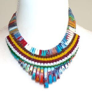 May include: A colorful, multi-layered, geometric statement necklace made of resin. The necklace is composed of rectangular pieces of resin in shades of blue, green, red, white, and yellow. The pieces are arranged in a fan shape, creating a unique and eye-catching design.
