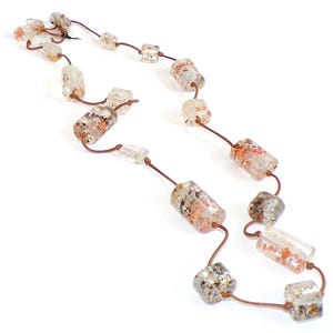 May include: A necklace featuring clear, cylindrical beads with reddish-brown and gold inclusions, strung on a brown cord. The beads are interspersed with smaller, square-shaped clear beads. The necklace is displayed against a white background.