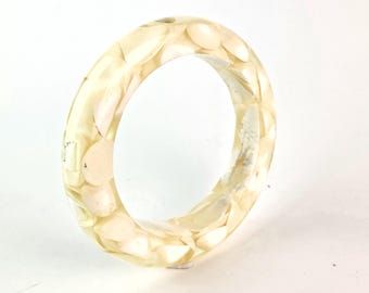 Könnte beinhalten: Ein klarer Armreif aus Harz mit eingebetteten, cremefarbenen, muschelartigen Stücken. Das Armband hat eine runde Form. Die Muschelteile sind von unterschiedlicher Größe und Form und erzeugen ein strukturiertes Aussehen.