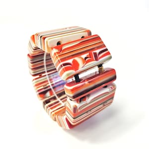 May include: A chunky bracelet with rectangular links. The links are multi-colored with stripes of red, orange, white, and brown. The bracelet is made of a glossy material and is set against a white background.