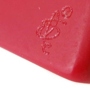May include: Close-up of a red object with a stylized, embossed design. The design appears to be a musical note or a stylized letter. The object has a smooth, glossy surface.