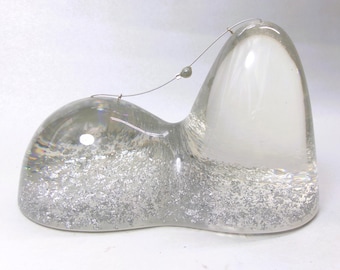 May include: A clear, smooth glass sculpture with a dual-peak, organic shape. The base is filled with dense, shimmering silver metallic flakes. A thin, curved silver wire arches between the two peaks, featuring a single small, round bead.