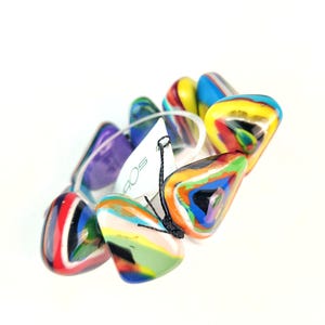 May include: A colorful bracelet featuring triangular, marbled beads in shades of red, blue, yellow, green, and purple. The beads are strung together with a clear elastic band. A white tag with black text is attached.