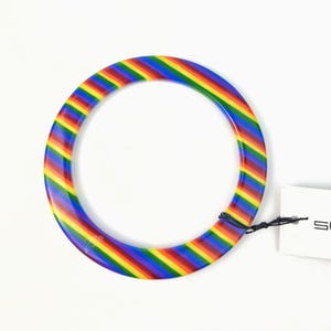 Puede incluir: Un brazalete a rayas de arcoíris. El brazalete tiene forma circular y presenta una vibrante gama de colores, incluyendo rojo, naranja, amarillo, verde, azul y morado. El brazalete parece estar hecho de un material brillante.