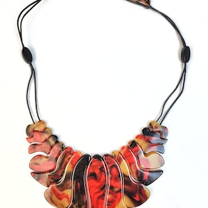 May include: A statement necklace with a large, abstract pendant made of black, red, and gold marbled plastic. The pendant is shaped like a series of overlapping curves and is attached to a black cord with a black clasp.