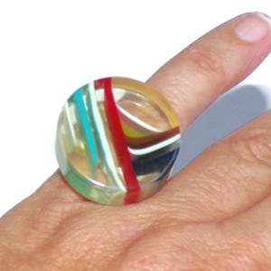 May include: A large, colorful ring with a clear, rounded design. The ring features vertical stripes in red, teal, white, and yellow. The ring is worn on a finger.