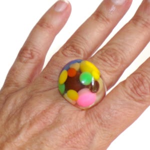 May include: A clear resin ring featuring an assortment of embedded colorful circular shapes in shades of yellow, pink, green, orange, and brown. The ring has a smooth, rounded, and polished surface, showcasing the multi-colored elements inside.