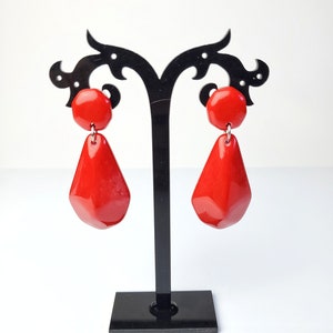 May include: A pair of red dangle earrings with a faceted teardrop shape. The earrings are displayed on a black earring stand.