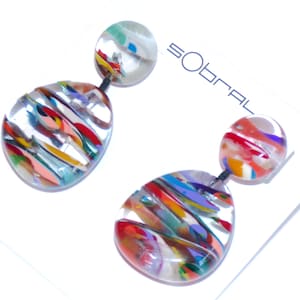 May include: A pair of colorful, abstract earrings. Each earring features a clear, teardrop-shaped pendant with vibrant streaks of red, orange, yellow, blue, and green. A smaller, round, colorful bead tops each pendant. The word "Sobral" is visible.