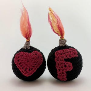May include: Two handmade crochet ornaments shaped like bombs. One features a red heart, the other a red letter "F". Each has a gray fuse with a flame of orange and yellow yarn. The ornaments are black.