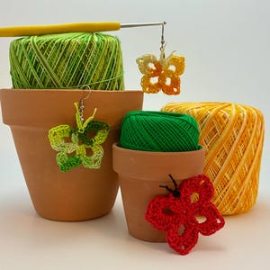 May include: A collection of crochet butterfly earrings in shades of green, yellow, and red, displayed with spools of yarn and a crochet hook. The yarn is in shades of green and yellow, and the earrings are hanging from a hook.