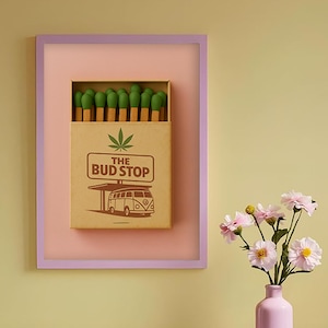 Op de afbeelding: Een ingelijst luciferdoosje kunstwerk in roze en paarse tinten. Het bruine luciferdoosje heeft de tekst "THE BUD STOP" en een cannabisblad. Groene lucifers zijn zichtbaar. Een roze vaas bevat roze bloemen.