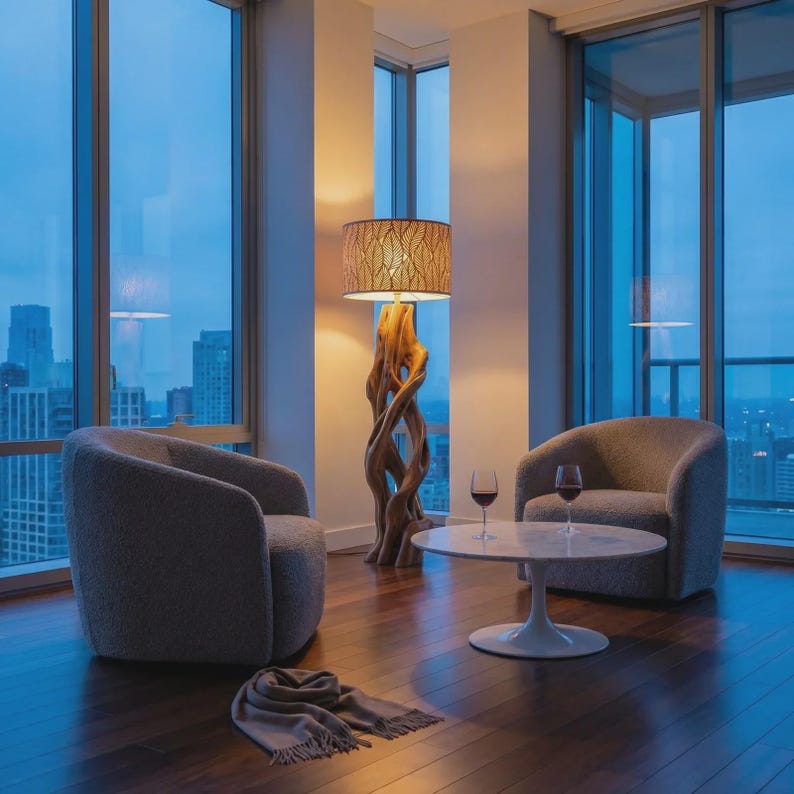 May include: A floor lamp with a twisted wooden base and a patterned drum shade, two gray upholstered armchairs, a white round pedestal coffee table with two glasses of red wine, and a gray fringed throw blanket on a dark wood floor.