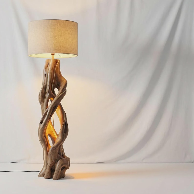 May include: A floor lamp featuring a base crafted from a single piece of natural, light-brown wood with a twisted, organic shape and hollow center. The lamp is topped with a cylindrical, beige fabric lampshade that emits a warm, soft glow.