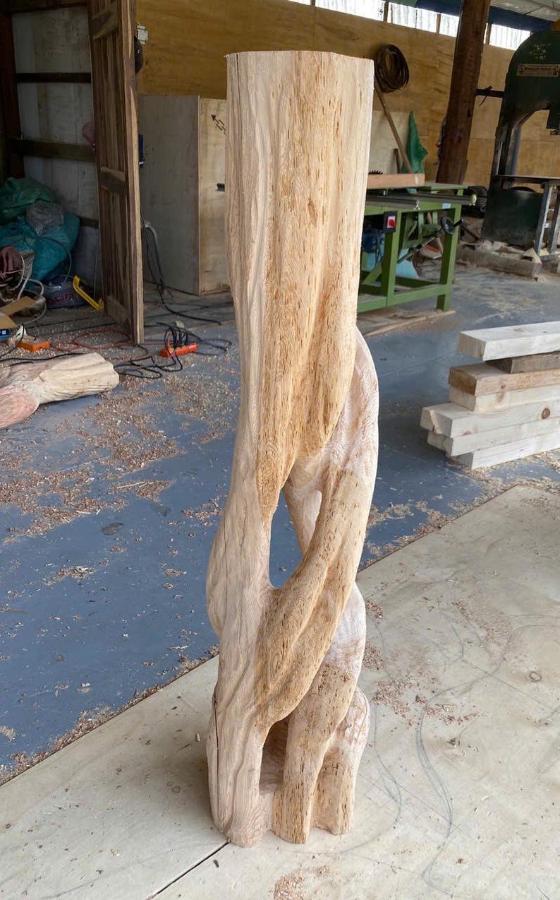 May include: A tall, light-colored wooden sculpture carved from a single log, featuring a vertical, twisted, and intertwined design with a hollow center. The surface displays natural wood grain patterns, textures, and light tan hues throughout the structure.