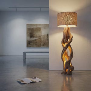 May include: A floor lamp featuring a base crafted from a twisted, natural light-brown wood trunk with an internal glow. The cylindrical lampshade is made of a tan material with a repeating cut-out pattern of detailed, veined leaves.