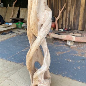 May include: A tall, light-colored wooden sculpture featuring a natural, twisted grain pattern. The structure consists of multiple intertwined, organic vertical forms that create open, hollow spaces throughout the center of the carved wood piece.