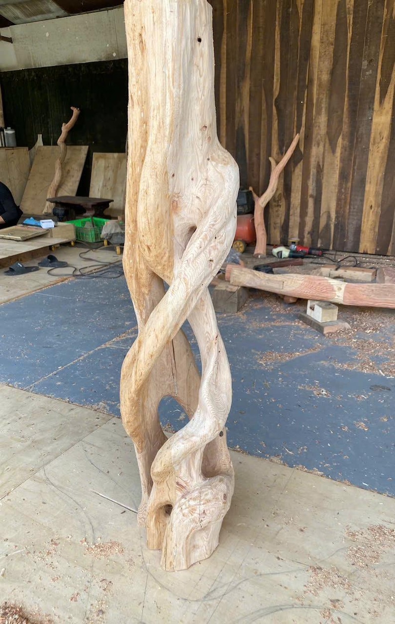 May include: A tall, light-colored wooden sculpture featuring a natural, twisted grain pattern. The structure consists of multiple intertwined, organic vertical forms that create open, hollow spaces throughout the center of the carved wood piece.