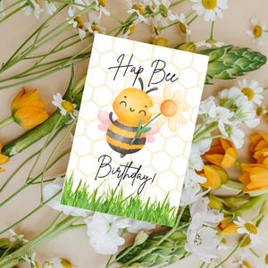 May include: A greeting card with a watercolor illustration of a smiling bee holding a flower. The card has the text "Hap Bee Birthday!" on a white background with a honeycomb pattern and green grass at the bottom. The card is surrounded by flowers.