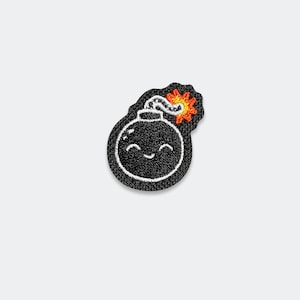 May include: An embroidered patch featuring a black spherical bomb with a smiling face, white stitched highlights, and a lit fuse with orange and yellow flames. The design is outlined in black thread with a textured, dense embroidery finish throughout.