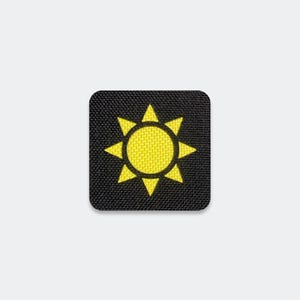 May include: A square-shaped patch featuring a black fabric background with a bright yellow embroidered sun icon in the center. The sun design consists of a solid circular core surrounded by eight triangular rays pointing outward in a star pattern.