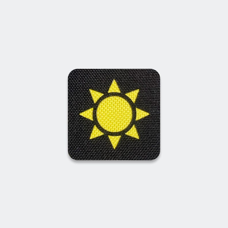 Puede incluir: Un parche de forma cuadrada con un fondo de tela negra y un icono de sol bordado en amarillo brillante en el centro. El dise&ntilde;o del sol consiste en un n&uacute;cleo circular s&oacute;lido rodeado por ocho rayos triangulares que apuntan hacia afuera.