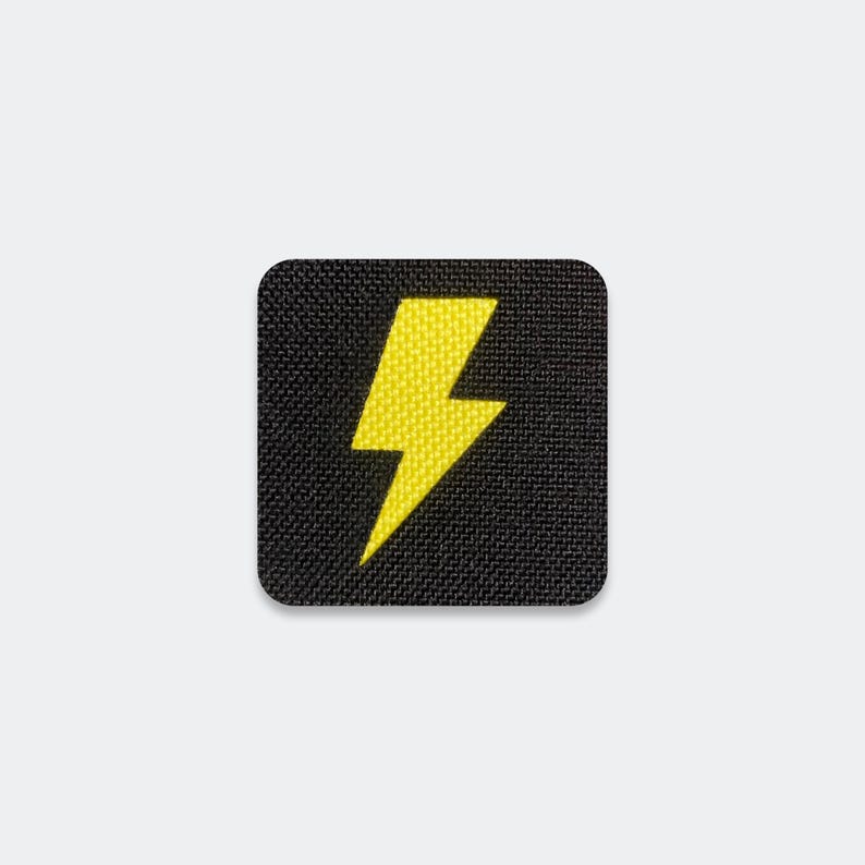May include: A square-shaped patch featuring a black fabric background with a textured weave. Centered on the patch is a bright yellow embroidered lightning bolt icon with sharp, angular edges and a clean, bold design.