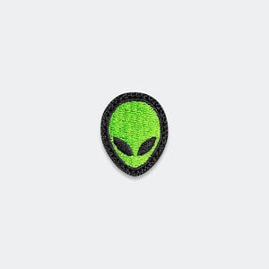 May include: An embroidered patch featuring a bright lime green alien head with large, almond-shaped black eyes. The design is enclosed within a thick, textured black border, creating a distinct contrast against the vibrant green center.