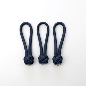 May include: Three dark navy blue zipper pulls, each featuring a braided cord loop and a spherical knot at the base. The textured, woven material forms a consistent, uniform shape for each of the three individual zipper pull attachments.