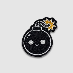 May include: Embroidered patch featuring a smiling cartoon bomb. The black bomb has white eyes and a curved mouth. A yellow explosion and a gray fuse complete the design. The patch is outlined in white.