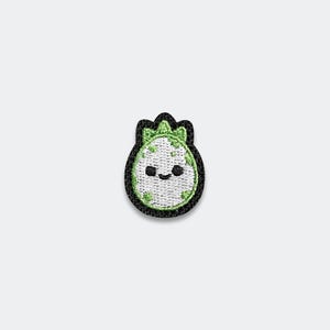 May include: An embroidered patch shaped like a rounded egg with a black border. The center is white with light green spots and a small green sprout at the top. It features a simple black stitched face with two small eyes and a curved mouth.
