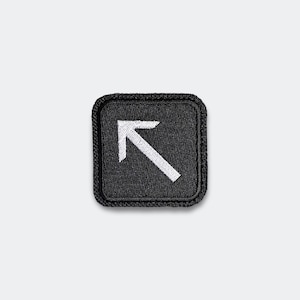 May include: A square black patch with a white arrow pointing up and to the right. The arrow is centered within the patch, which has a stitched border. The patch is likely designed to be sewn or ironed onto clothing or accessories.